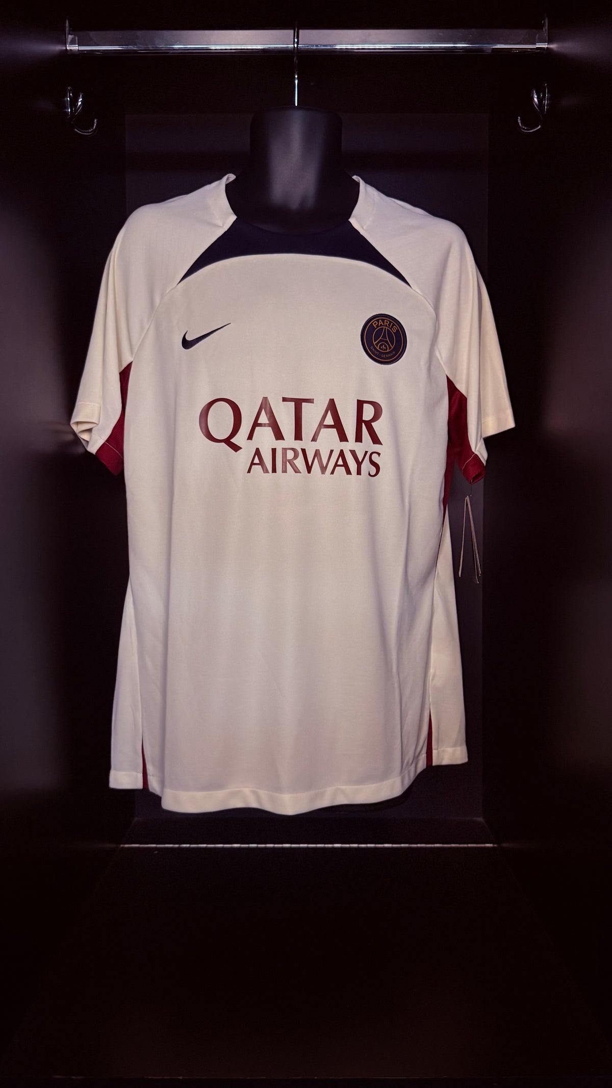 A white Paris Saint-Germain soccer jersey with 'QATAR AIRWAYS' sponsor text, displayed on a black mannequin torso against a dark background.